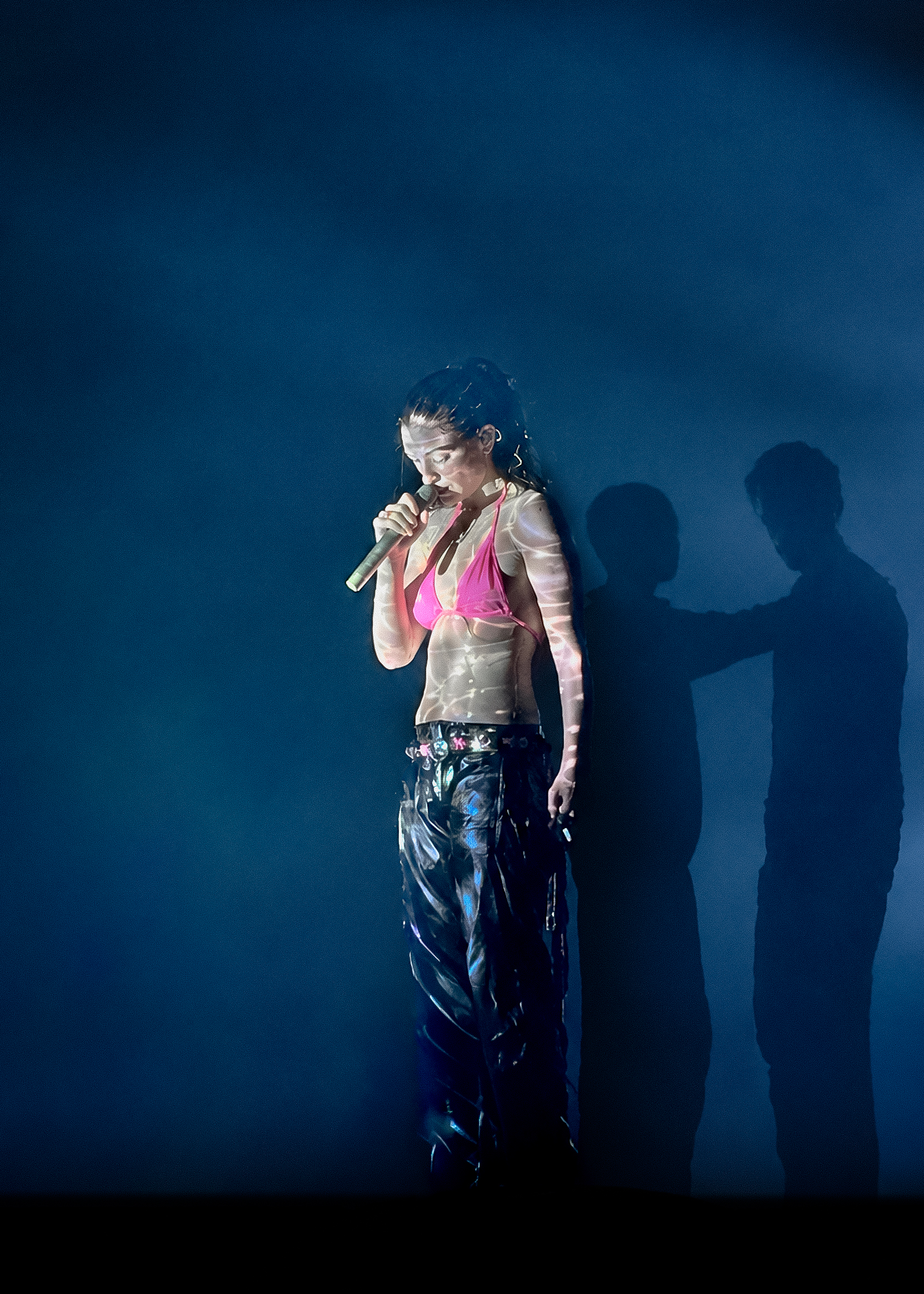 A singer holding a microphone under blue stage lighting, with projected light and shadows behind her.